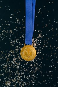 a gold metal. Photo by Nataliya Vaitkevich: https://www.pexels.com/photo/close-up-shot-of-a-gold-medal-on-a-black-surface-6120397/